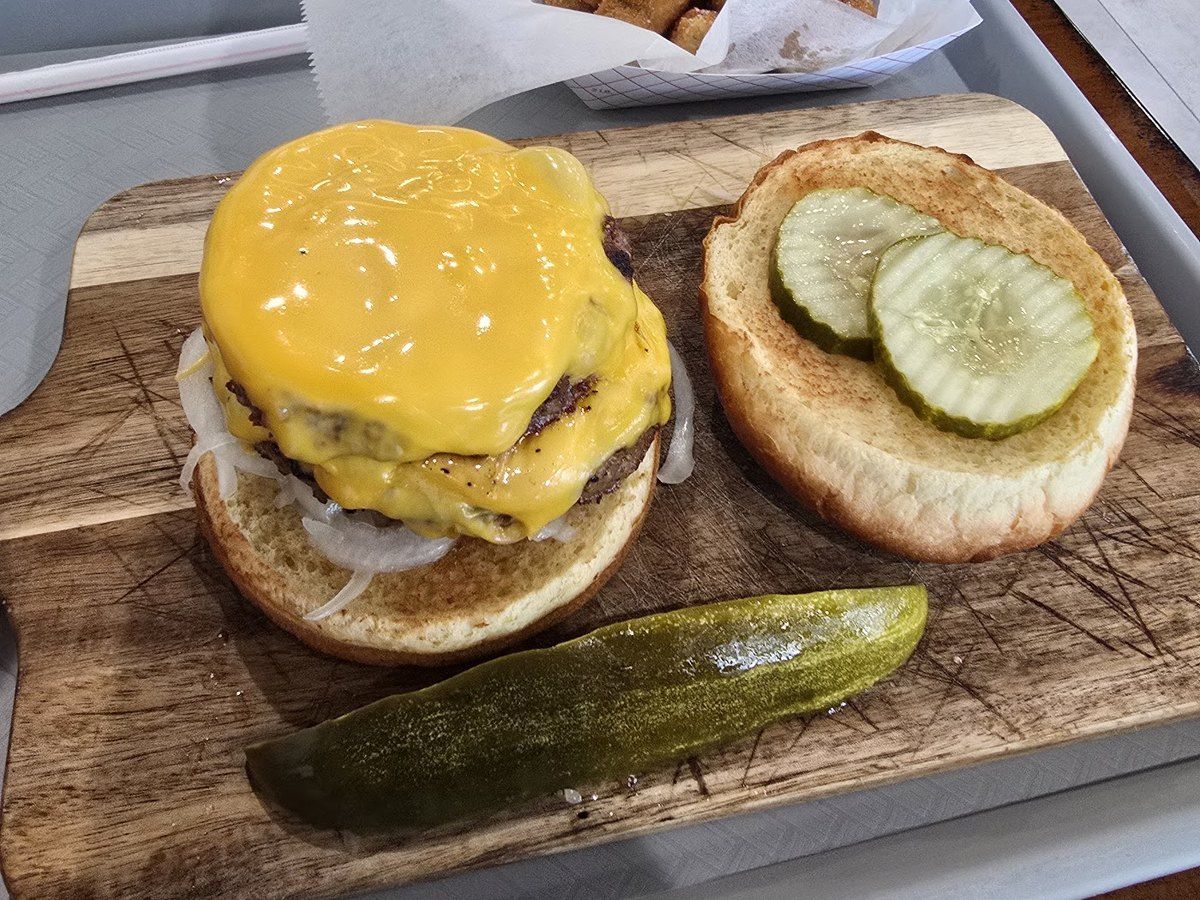 A double cheeseburger on a wooden board with a pickle spear