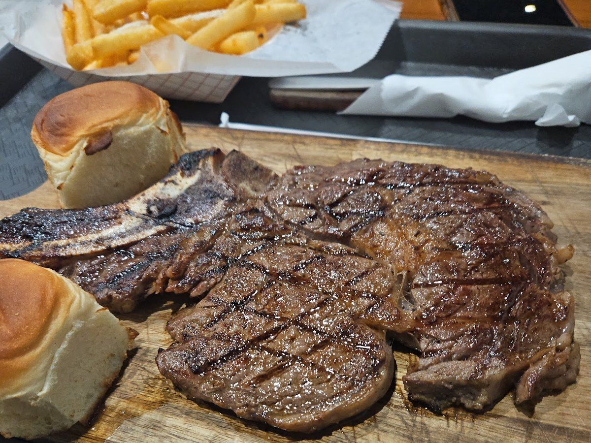 A grilled T-bone steak on a wooden board with Hawaiian rolls and fries
